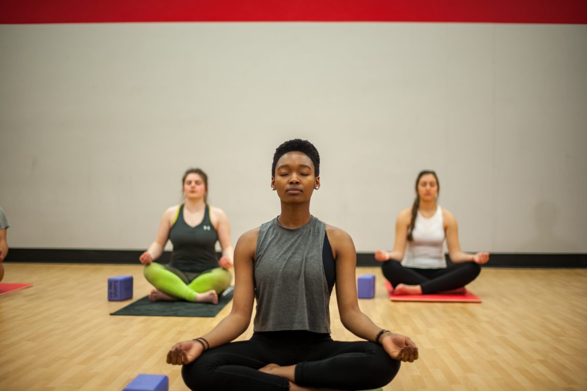 Photo of three fitness instructors sitting cross-legged on the ground with their eyes closed performing meditation in an indoor fitness studio.