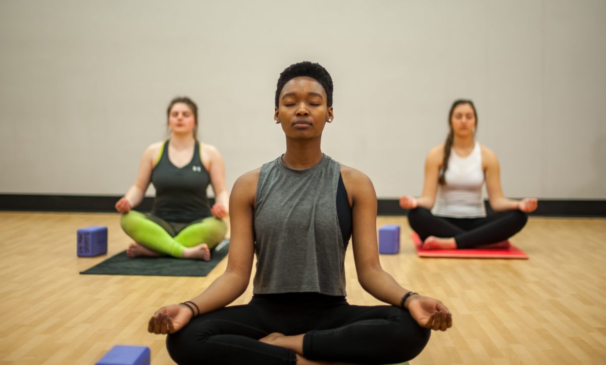 Photo of three fitness instructors sitting cross-legged on the ground with their eyes closed performing meditation in an indoor fitness studio.