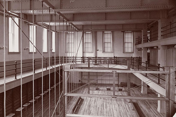 Image inside of the ladies hall gym from 1900. Wood floors and a second story overlooking the first highlight the image.