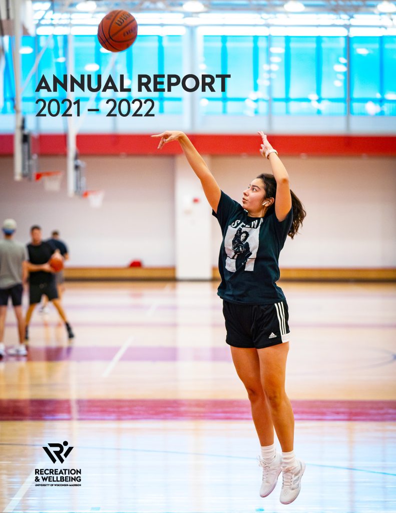 This is the cover of our 2021-22 Annual Report. It features "Annual Report 2021-22" in the upper left corner and our Rec Well logo in all black in the lower right corner. The image underneath shows a woman shooting a basketball into a hoop at the Nick. She is mid-shoot and has both feet off the floor.